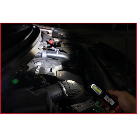 Een hand houdt een LED-werklamp in een donkere motorruimte. De lamp verlicht motoronderdelen.